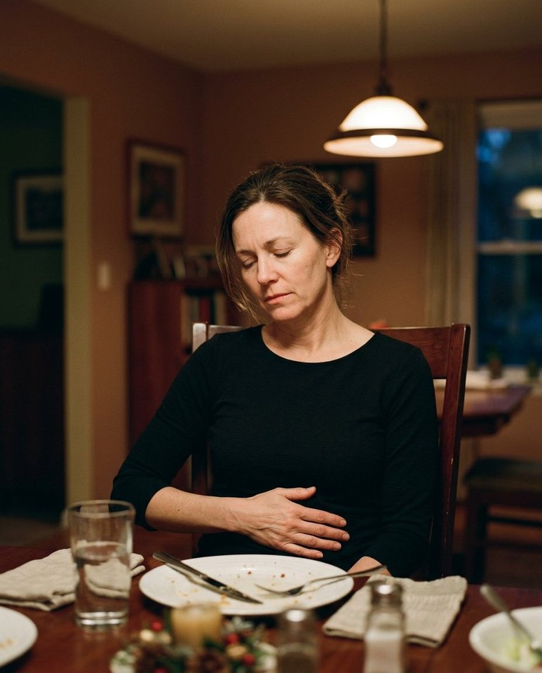 A woman at her dining room table after dinner, hand resting on her midsection, eyes closed
